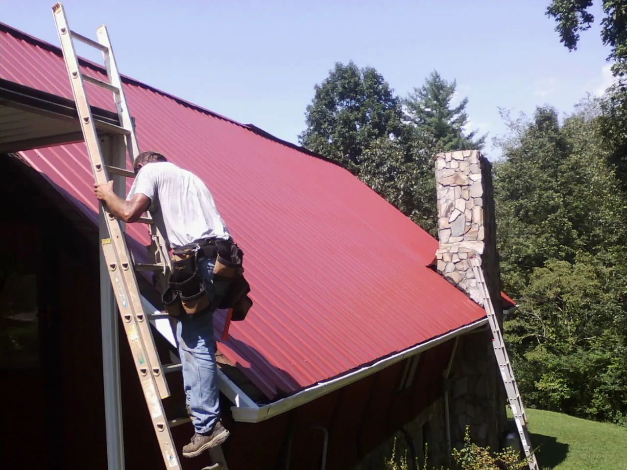 Roofer on a ladder beside a red metal roof and stone chimney — Soffit & Fascia Repair by Roofing Brothers in Worth